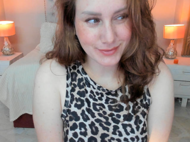 KatyaRogersX with wavy brown hair, smiling, wearing a leopard print sleeveless top, showing her pale skin and subtle freckles, sitting in a softly lit bedroom.