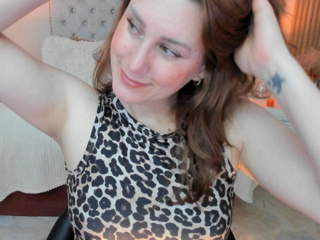 KatyaRogersX with arms raised, wearing a leopard print top, has red lipstick, brown wavy hair, and a star tattoo on her right forearm, smiling at the camera.