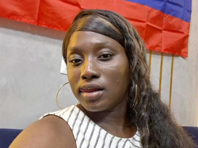 CocoaBell, with dark skin and shiny brown hair in a braid, wearing a striped sleeveless top and large hoop earrings, looking slightly to the side with a subtle smile.