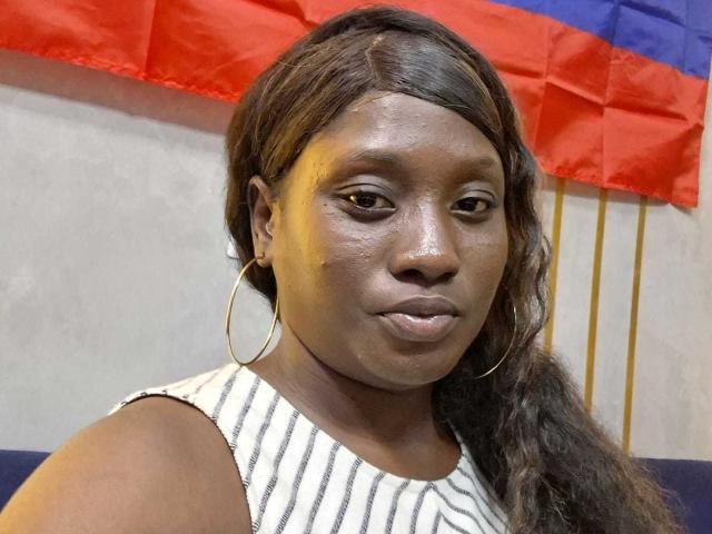 CocoaBell has dark brown skin, long wavy hair, and wears large hoop earrings with a white and black striped sleeveless top, looking serious with subtle makeup.