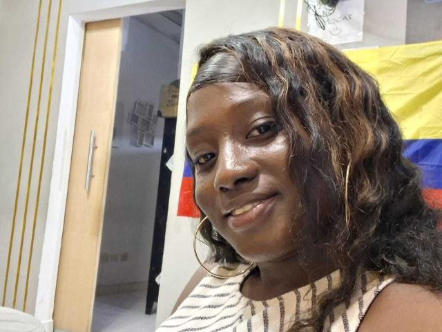 CocoaBell has dark brown skin, wavy brown hair with highlights, and is smiling with her small tits visible. She's wearing a striped sleeveless top and large hoop earrings.
