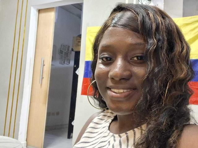 CocoaBell with shiny dark brown skin, wavy black hair, wearing a zebra-striped top, hoop earrings, smiling, showing teeth, standing in a room with a Colombian flag.