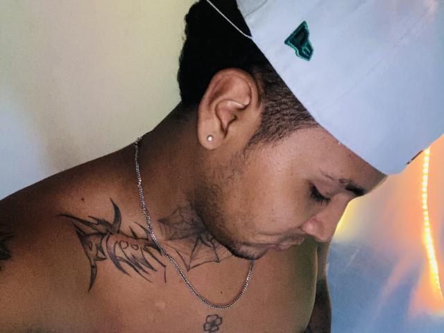 AxellKing69 with dark skin, short curly hair, white cap, small earrings, chain necklace, tattoos on shoulder and neck, looking down, showing off a clean-shaven face and a slight beard.