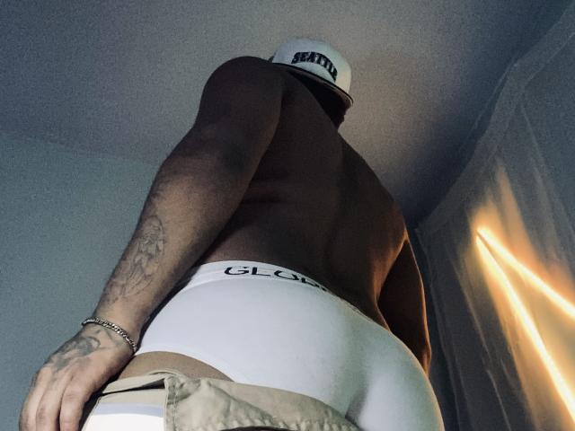 AxellKing69, muskulös, ohne Hemd, trägt weiße "GLOOM"-Unterwäsche, eine Mütze und ein Armband, mit dem Rücken zur Kamera, sichtliches Tattoo am rechten Arm, beige Hose, die teilweise heruntergezogen ist, Neonlicht auf der rechten Seite..