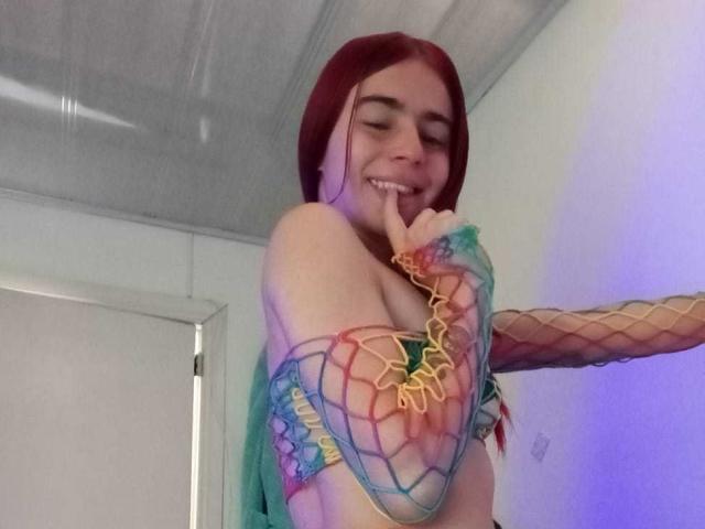 LoveMaxim mit roten Haaren, lächelnd, mit Finger im Mund, trägt ein farbiges Netzoberteil, das ihre Brüste zeigt, und hat einen Arm mit Regenbogenmuster, in einem hellen Raum..