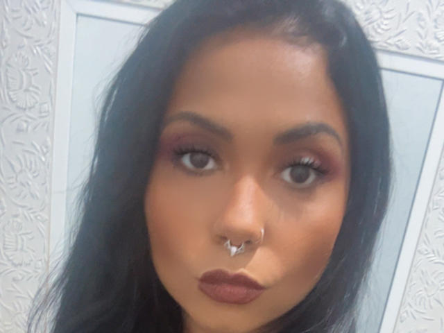 Karollz with tan skin, dark hair, brown eyes, and a septum piercing, wearing red eyeliner and brown lipstick, looking straight at the camera with a pouty expression.