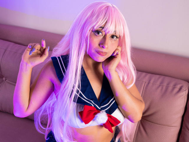 AilenRosse with pink hair, wearing a navy blue and white sailor outfit with a red bow and white pom-pom, sitting on a pink leather couch, hands near face.