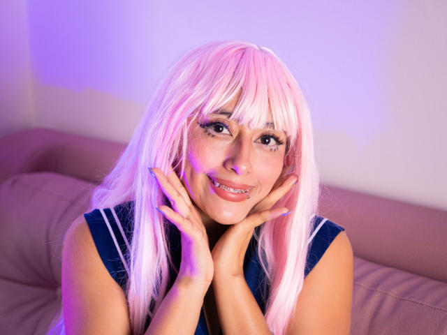 AilenRosse with pink hair, smiling, hands on cheeks, wearing a blue top, purple and pink lighting, visible teeth, long eyelashes, light skin, and pink nails.