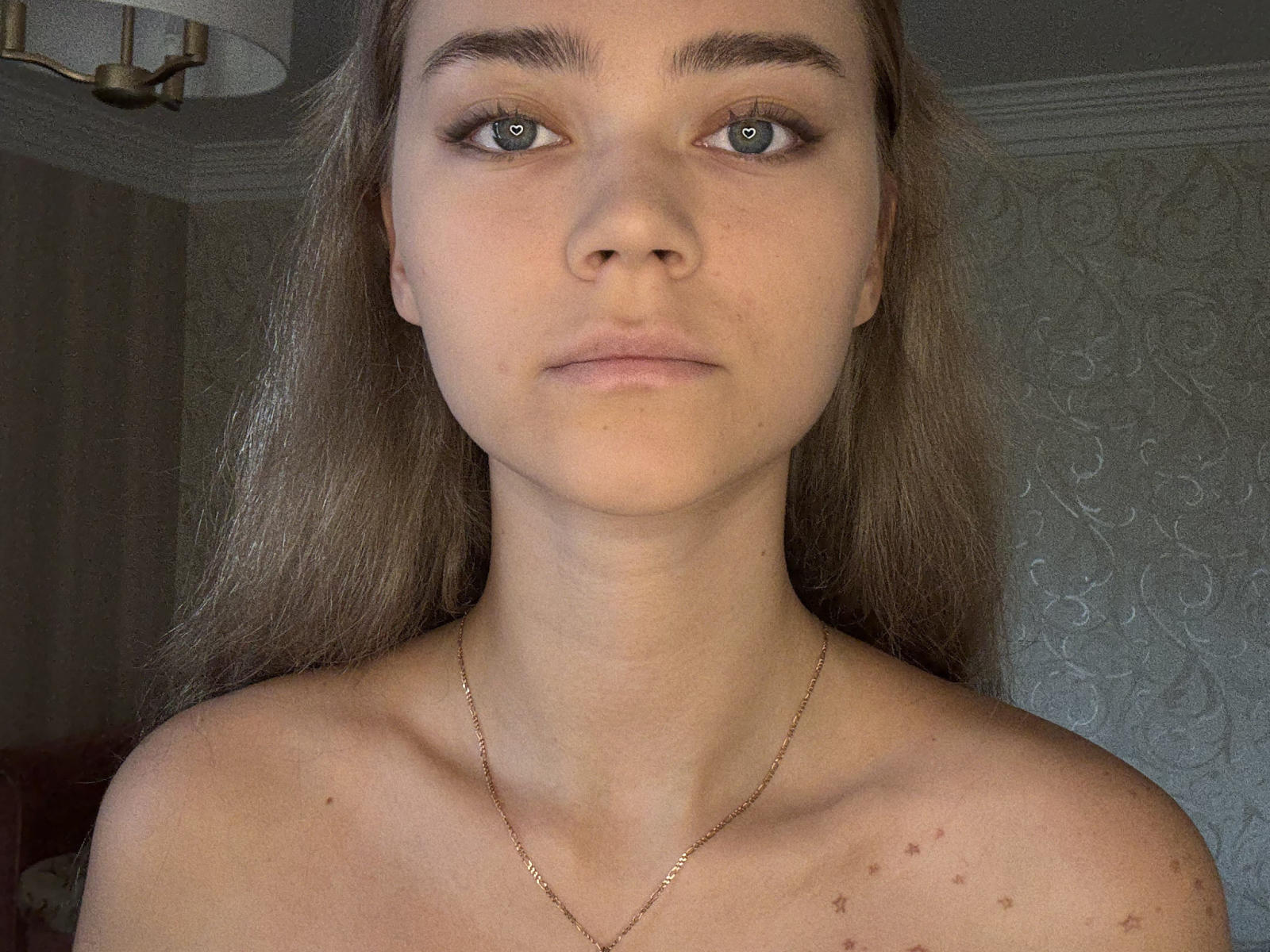 Thomasina Ortz tiene la piel clara, ojos verdes y cabello largo castaño, lleva un collar dorado, tiene pequeñas pecas en la parte superior del pecho y mira directamente a la cámara con una expresión neutra..