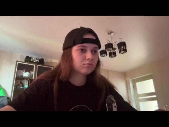 RosemarieRacer with long brown hair, wearing a black baseball cap and black shirt, has a serious, slightly annoyed expression. She's in a room with a ceiling fan.