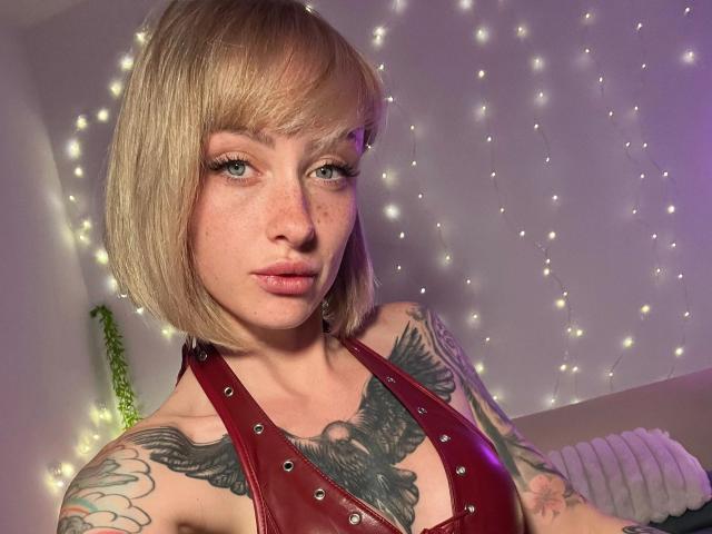 HaileyhopeXXX-hot with blonde bob, green eyes, and freckles, wearing a red leather halter top, showcasing tattoos on her chest and arm, surrounded by fairy lights.