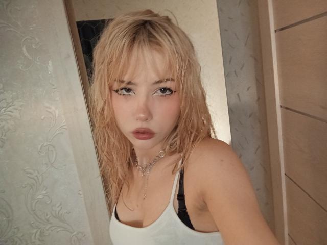 Blonde EileneSzala with wet hair, white tank top, black bra straps, chain necklace, and pouty lips stares into camera. Boobs slightly visible, arms at sides.