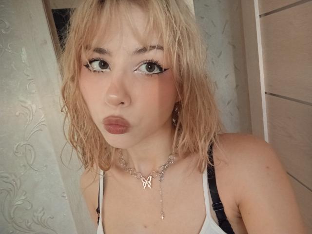 Blonde EileneSzala with wet hair, pouty lips, and thick eyeliner, wearing a white tank top and butterfly necklace, has small tits and a black strap over her shoulder.