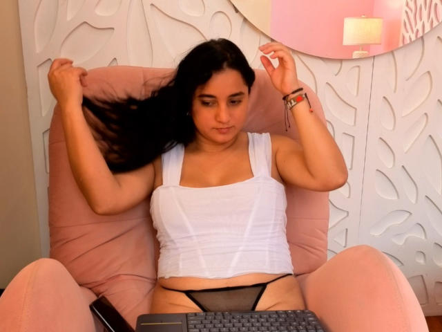 LisaStarkm with long black hair, white tank top, black mesh panties, pink chair, holding hair, laptop in front, pink wristband, white wall with leaf patterns, relaxed expression.