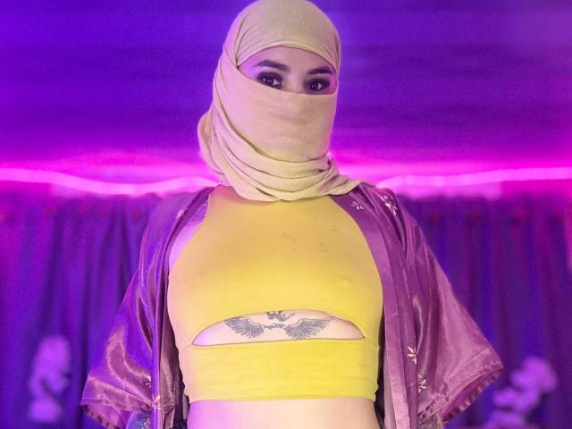 MalakCote wearing a yellow crop top with a cutout over her tits, a pink shiny jacket, and a beige hijab with her face covered, purple neon lights in background.
