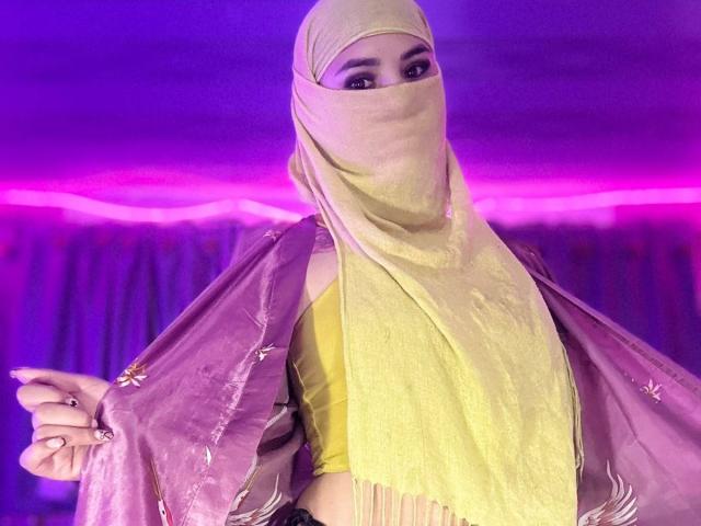 MalakCote with a yellow and pink hijab, yellow top, and pink scarf, holding her scarf open, showing her face and upper body, with purple lighting.