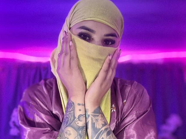 MalakCote with yellow mask, hands covering face, pink nails, tattooed arms, purple and pink lighting, wearing shiny pink jacket, yellow headscarf, dark eyebrows, looking at camera.