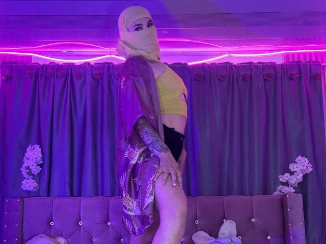 MalakCote with blonde hijab, yellow sports bra, black lace panties, and purple sheer robe, standing, showing tattooed arm, purple neon lights, and floral decor in background.