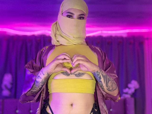 MalakCote with yellow hijab, yellow crop top, and purple robe, showing heart hands, tattooed arms, and exposed chest with small tits and butterfly tattoo. Purple neon light in background.