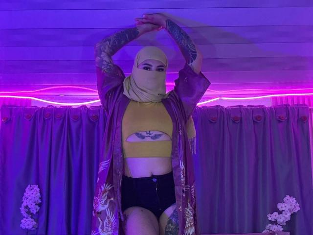 MalakCote with tattooed arms, yellow crop top, black shorts, purple robe, yellow mask, and black underwear, arms raised, purple LED strip lighting, floral curtain.