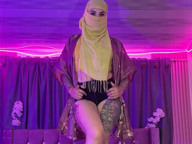 MalakCote with a yellow hijab, purple shawl, black fringed shorts, and tattoos, sitting on a couch, legs crossed, under purple LED lights, looking at the camera.