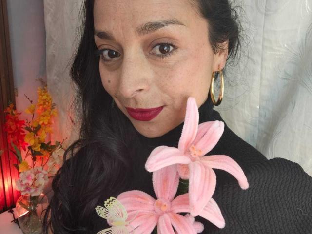 StangelarRivera, avec de longs cheveux noirs, portant du rouge à lèvres, des boucles d'oreilles dorées et un pull noir à col roulé, tenant des fleurs en tissu rose et une décoration de papillon..