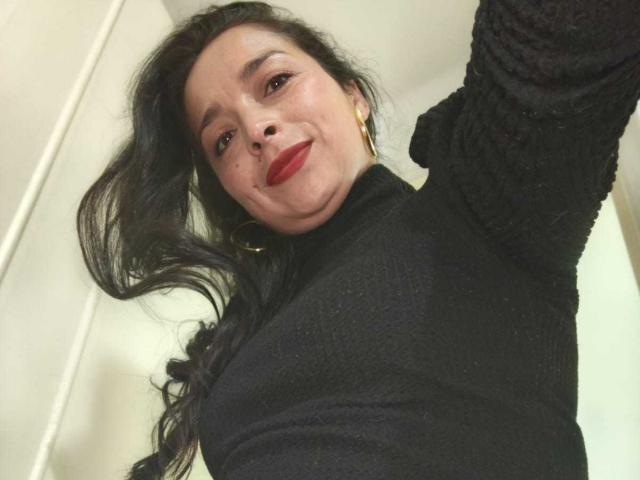StangelarRivera, con cabello largo y negro, vestida con un suéter negro texturizado, lápiz labial rojo y aretes dorados, sonriendo a la cámara y levantando su brazo. Sus senos no son visibles..