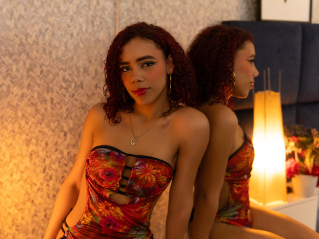 ValeryMartinez with curly red hair, wearing a colorful strapless dress, gold hoop earrings, and a necklace, sitting in front of a mirror, reflection showing her ass. Warm light.