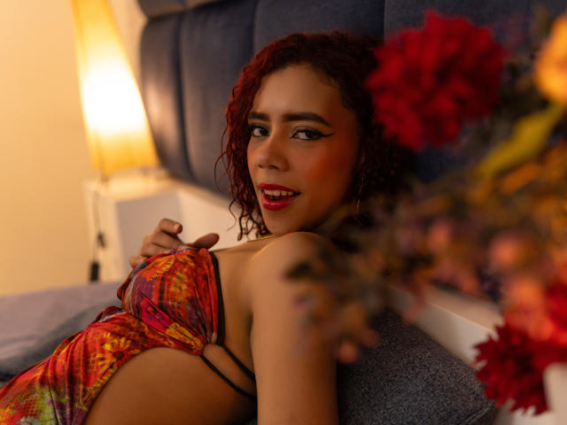 ValeryMartinez with curly red hair, wearing a colorful, tie-dye, strapless top, lying on a bed, looking at the camera, with red flowers in the foreground, smiling.