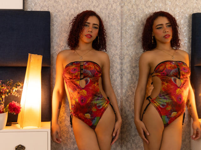 ValeryMartinez with curly red hair, wearing a colorful, strapless one-piece with cutouts, showing her ass and small tits, standing in front of a mirror, lit by a warm lamp.