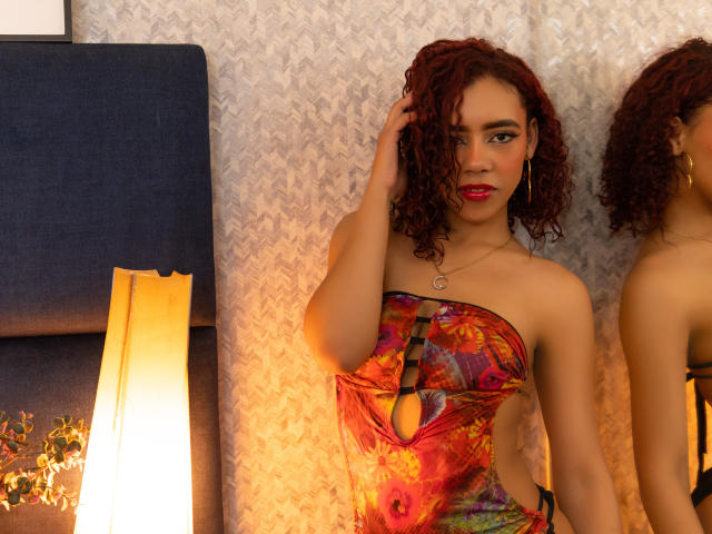 ValeryMartinez with curly red hair, wearing a colorful strapless dress, has her hand in her hair, red lipstick, gold earrings, and a necklace, standing in a warmly lit room.