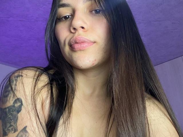 SophiaMarx with long brown hair, pouty pink lips, slight acne, and a tattoo on her left shoulder. She's topless, showing her bare shoulders and a small, visible nipple.
