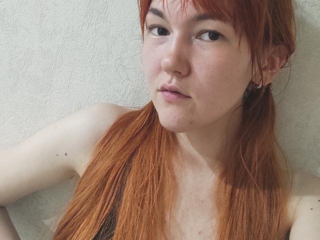 SheritaChieng a la peau pâle, des cheveux roux en couettes, de petites taches de rousseur, et elle regarde sur le côté avec une légère moue. Elle porte un débardeur noir..