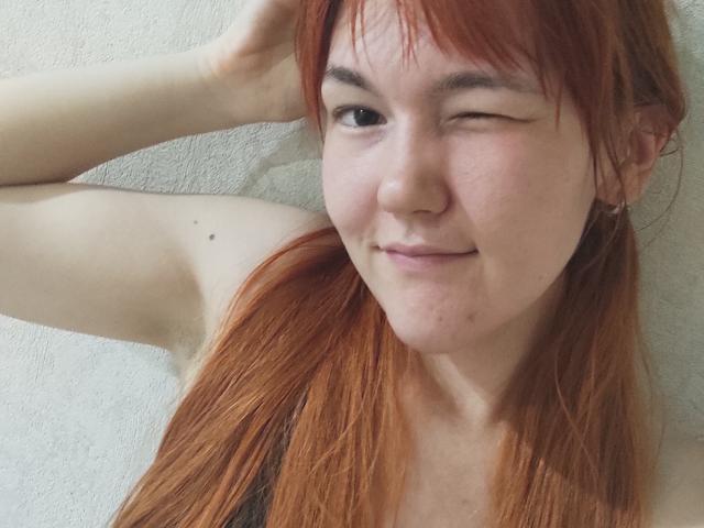 SheritaChieng with red hair, winking, smiling, showing her bare shoulder with a small mole, wearing a black tank top, against a white textured wall.