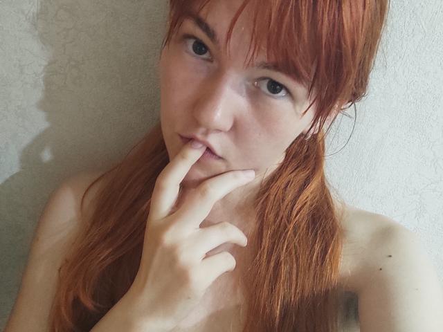 SheritaChieng has red hair, pale skin, and blue eyes, with her finger touching her slightly parted lips, looking seductive. She's topless, with visible bare shoulders.