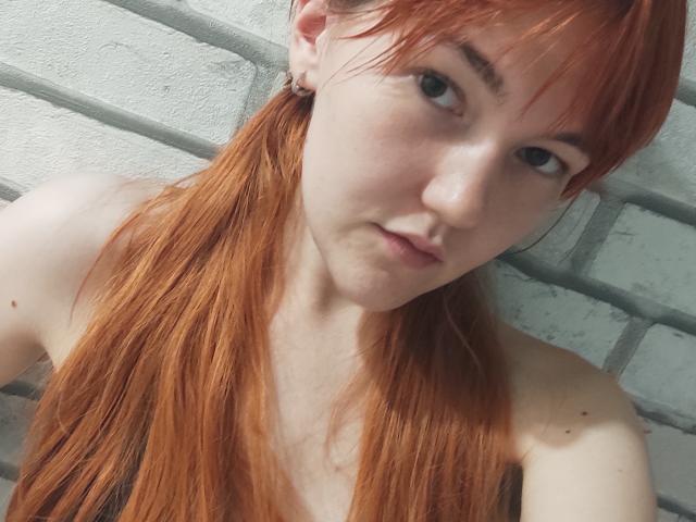 SheritaChieng with red hair, pale skin, small nose, and green eyes, looking at the camera with a slight smirk, wearing no top, showing her bare shoulders and upper chest.