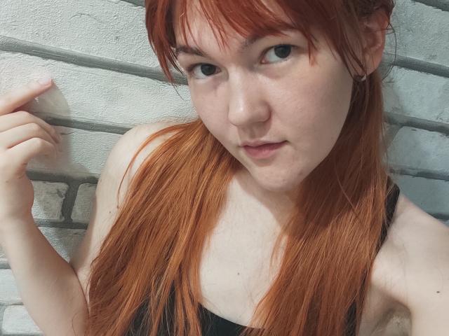 SheritaChieng with red hair, pale skin, and black tank top, holding a piece of clear plastic, looking slightly to the side with a subtle, confident smile.