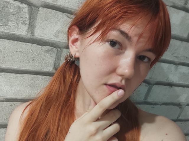 SheritaChieng has red pigtails, a cute face with freckles, and is biting her finger with a slight pout, wearing a leopard print earring, showing her bare shoulders.