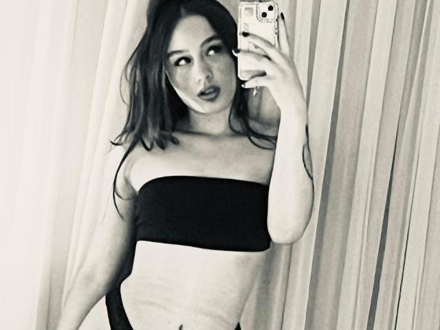 LucieDolly with dark hair, wearing a black strapless top and black underwear, taking a mirror selfie, showing her tits and ass, holding a phone with a white case.
