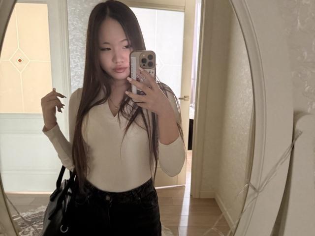 DirtyAsianLunachka com cabelos castanhos longos, blusa branca de manga comprida, calça preta de cintura alta, segurando o celular em um selfie no espelho, sorrindo, mão direita ligeiramente levantada, bolsa preta no lado esquerdo..