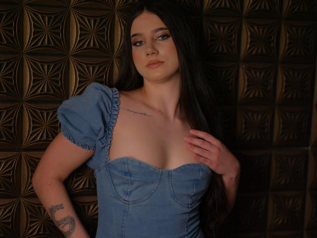 LexiSolene, avec de longs cheveux noirs, portant une robe bleue, touchant sa poitrine, montrant de petites seins, un tatouage sur le bras droit et un tatouage sur la poitrine gauche, debout devant un mur à motifs..
