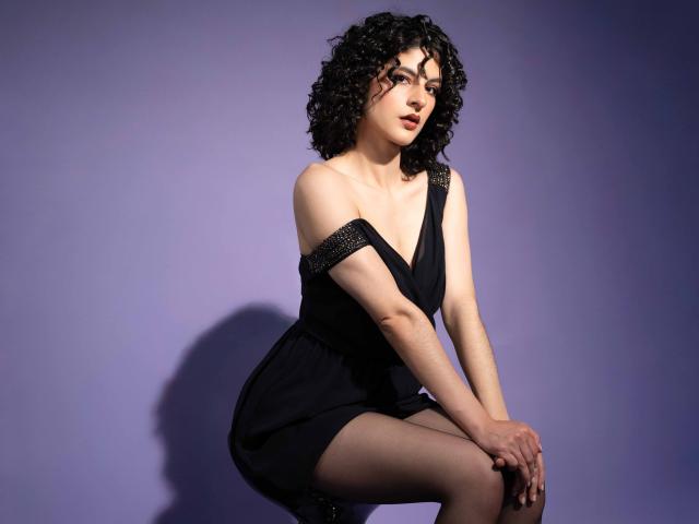 RoxyGraff, with curly black hair, wearing a black off-shoulder dress with sparkly straps, sitting with legs crossed, looking slightly to the side, on a purple background.