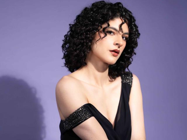 RoxyGraff with curly black hair, pale skin, and a serious expression, wearing a black, off-shoulder dress with silver studs, showing her tits and a subtle, confident smile.
