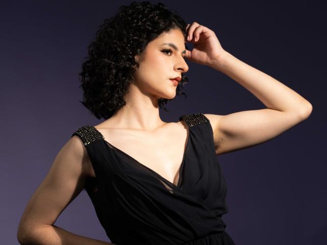 RoxyGraff with curly black hair, wearing a black dress with studded shoulders, raises her right arm, showing a muscular arm and armpit, against a dark purple background.