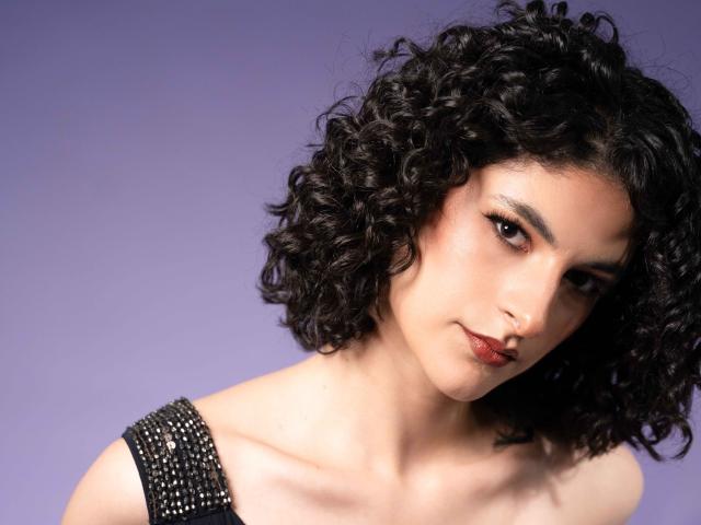 RoxyGraff has curly black hair, light brown skin, and dark red lipstick. She's wearing a black, beaded shoulder strap, looking slightly to the side with a subtle smile.