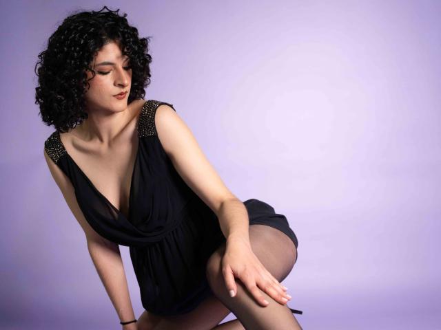 RoxyGraff with curly black hair, wearing a deep black dress with sparkly shoulder straps, bent forward, showing her tits, and holding her thigh, against a purple background.
