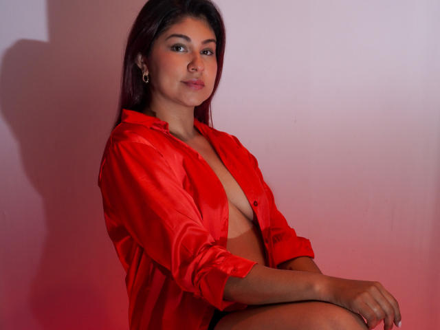 SamanthaBlossom with dark brown hair, wearing an unbuttoned red satin shirt, exposing her small tits, sitting with a confident smile, and a slight red light shadow on her.