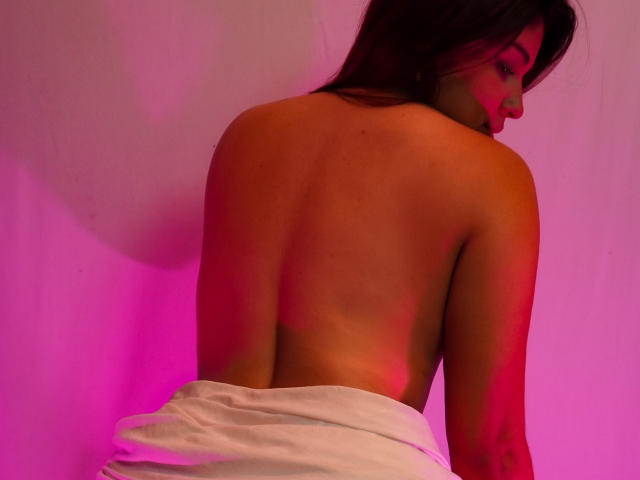 Le dos nu de SamanthaBlossom, avec ses cheveux bruns lâchés, portant une serviette blanche enroulée autour de sa partie inférieure, sous un éclairage rose et rouge. Elle regarde par-dessus son épaule..