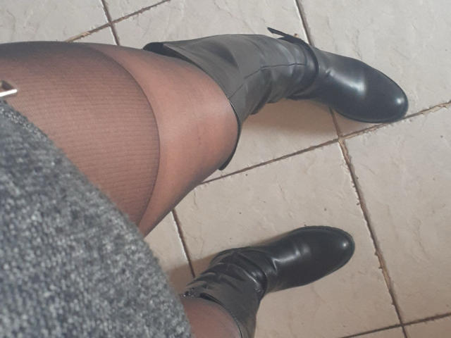Catiminiclan's legs in black sheer pantyhose, wearing black leather boots, with gray textured skirt, standing on beige tiled floor, photo angle showing from above.