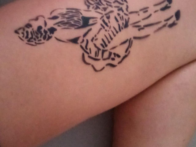 Tattoo am Arm von Catiminiclan, das ein schwarzes, stilisiertes Design eines Vogels mit ausgebreiteten Flügeln und einem gemusterten Schwanz zeigt, platziert knapp oberhalb ihres Knies..